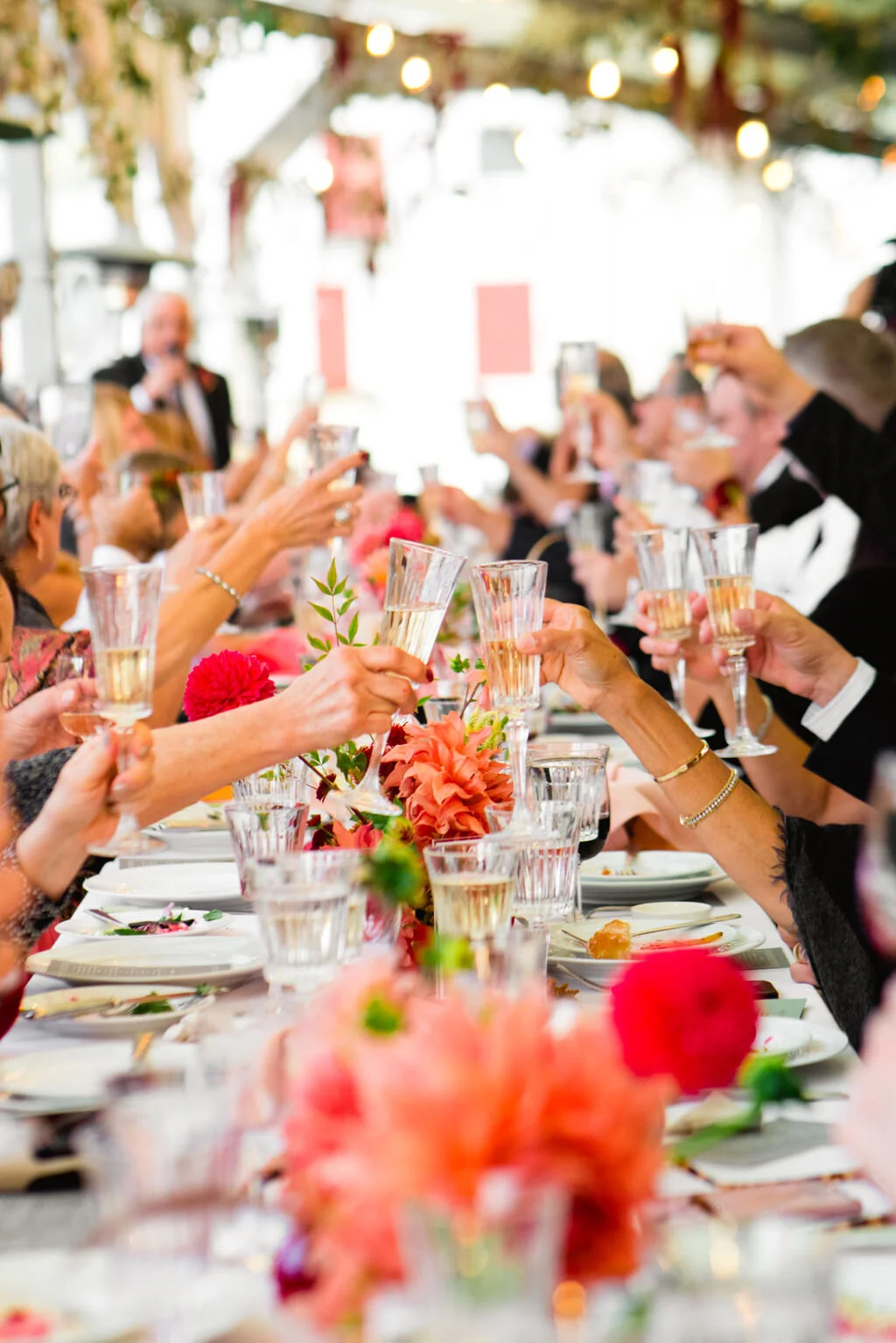People raising glasses in a toast at a formal celebration or wedding reception with a decorated table and floral centerpieces.
