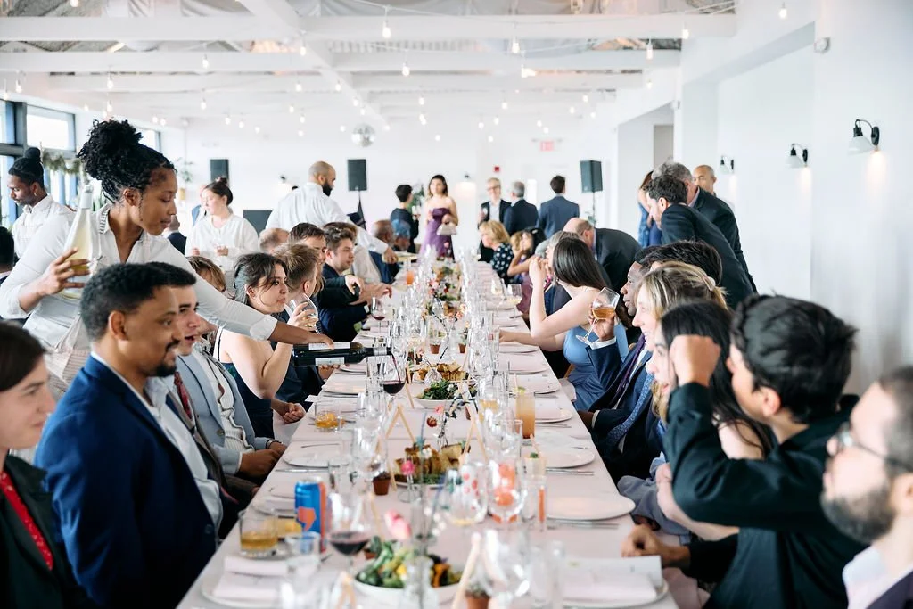 A diverse group of people in formal attire sitting at a long banquet table during a celebration or event with white walls, string lights, and waitstaff serving drinks.