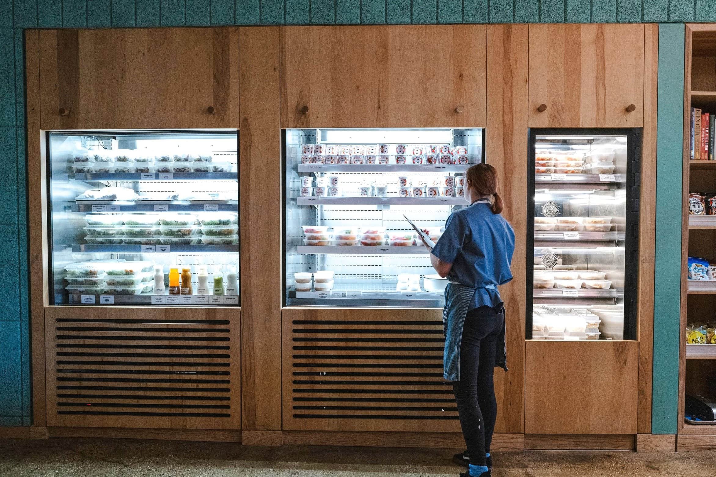 A woman in a blue shirt and black pants stands in front of refrigerated display cases filled with packaged food items, in a grocery store.