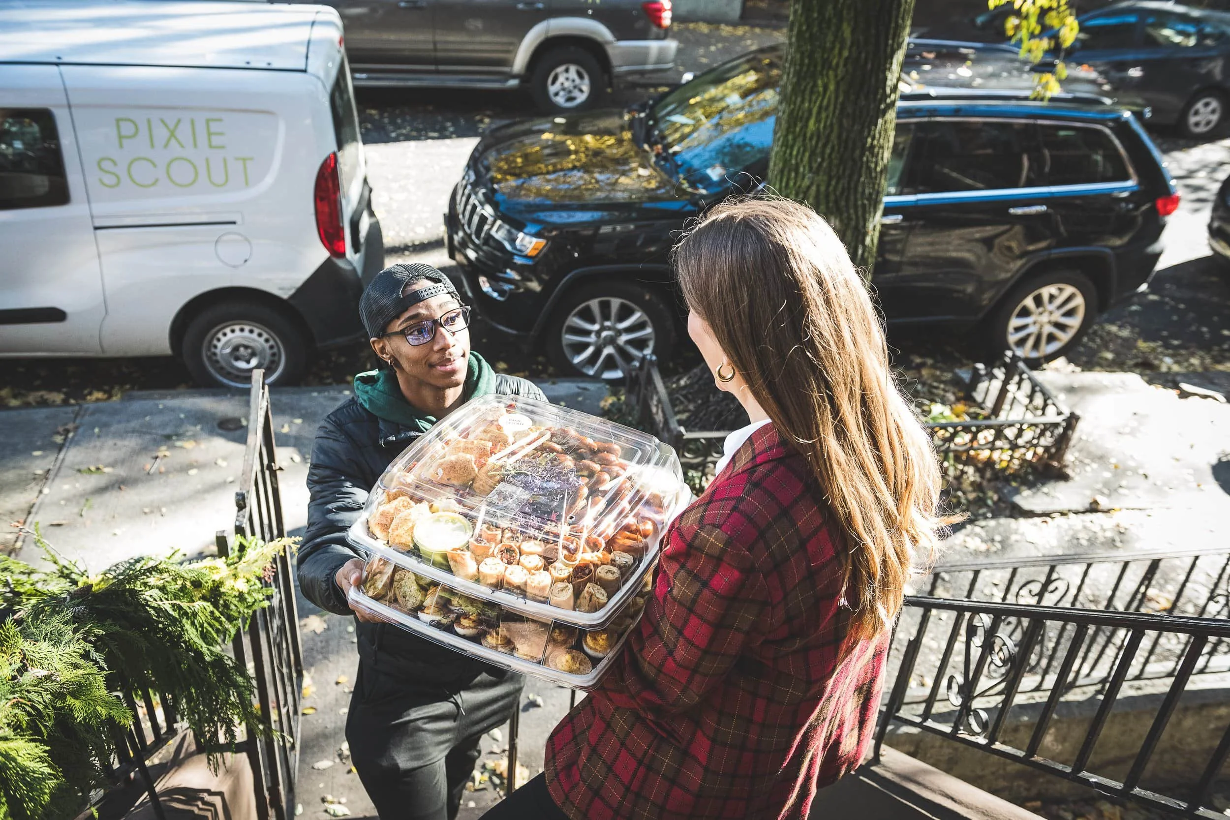 A woman in a red plaid jacket receiving a large clear container of assorted food from a woman in glasses and cap on a sidewalk with parked cars and fallen leaves in the background.