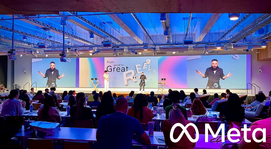 A conference or seminar with a large audience seated at tables, looking at a stage with three speakers. The stage has a large screen displaying the words 'From good to great' and colorful letters spelling 'FEELS.' The Meta logo is in the bottom right corner of the image.