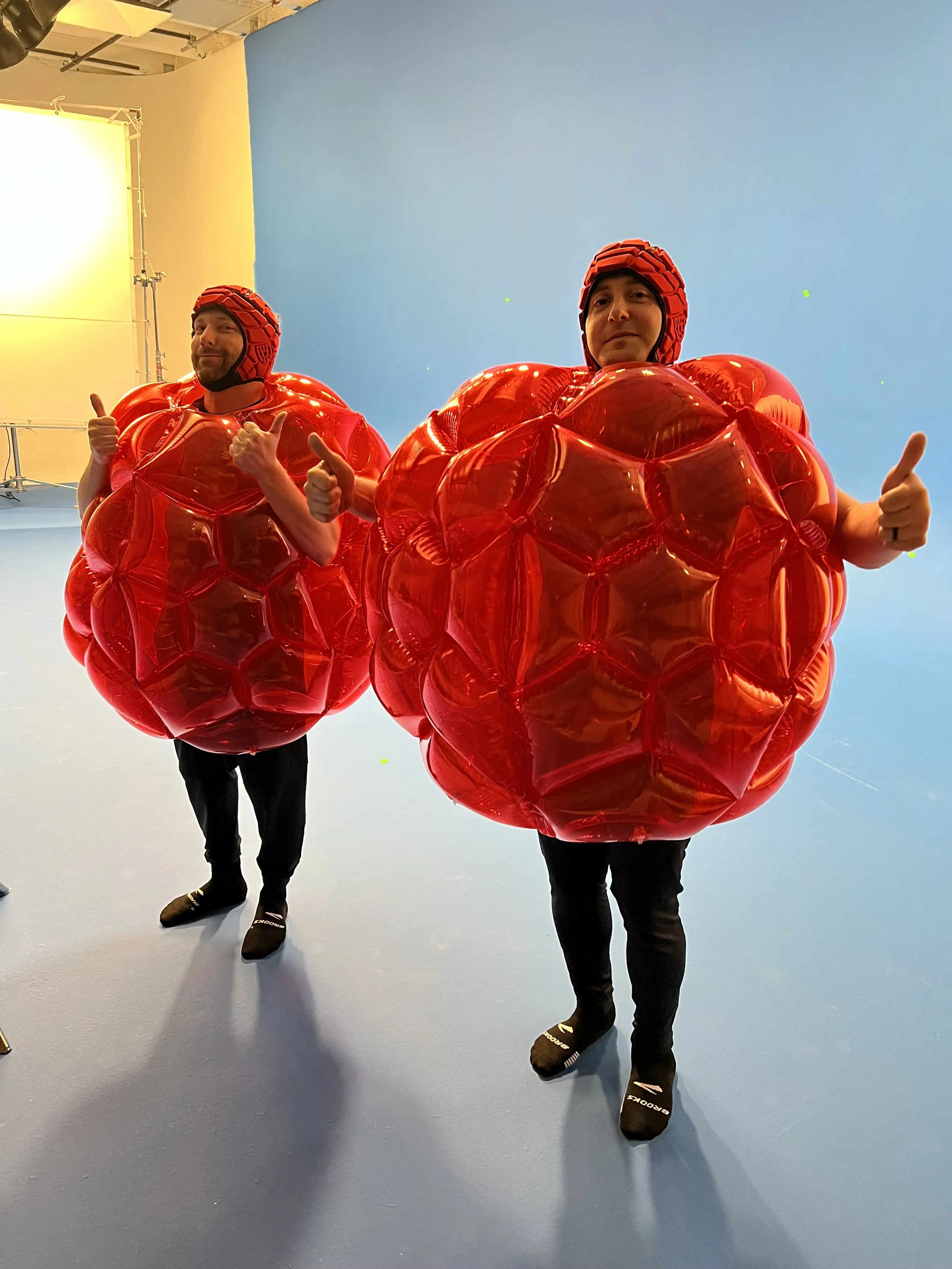 Two people dressed in inflatable red balloon costumes resembling large tomatoes, wearing helmets, standing indoors against a blue wall, and giving thumbs-up gestures.