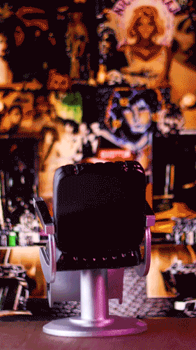 A black office chair in front of a cluttered desk with various items, including bottles, a computer, and decorative objects, with a colorful, busy background