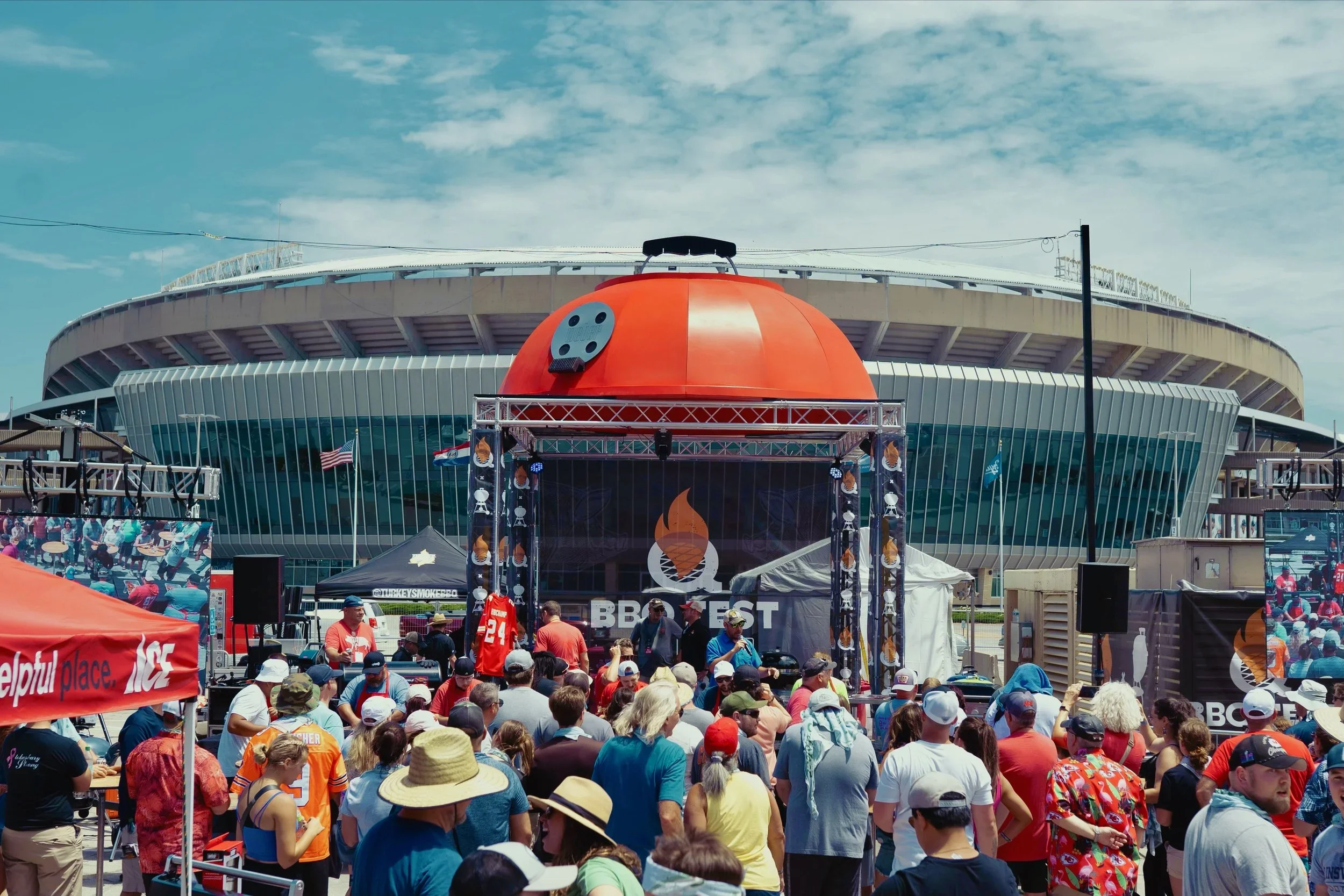 Crowd gathering at an outdoor event in front of a large stadium, with a food truck and stage with a BBQ fest logo, American flags, and various tents and displays.