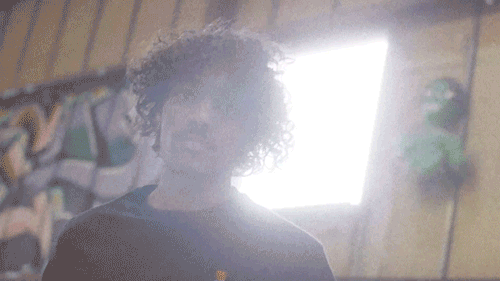 A person with curly hair standing indoors with wooden walls, a bright window behind, and colorful artwork on the wall.