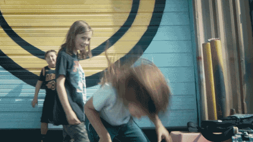 Three children playing indoors near a yellow and blue wall with a circular design, one girl with red hair bending over, another girl with long hair standing, and a third girl in the background with curly hair.