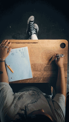 Overhead view of a person sitting at a wooden desk, writing in a notebook with a pen, with a pair of sneakers visible at the edge of the desk