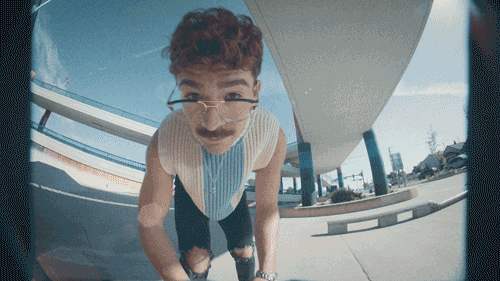 A young boy with glasses looking into a fisheye lens close-up, outdoors near a modern curved building.