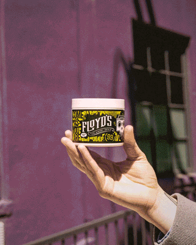 A hand holding a container with a yellow and black design labeled Floyd's against a purple wall background.