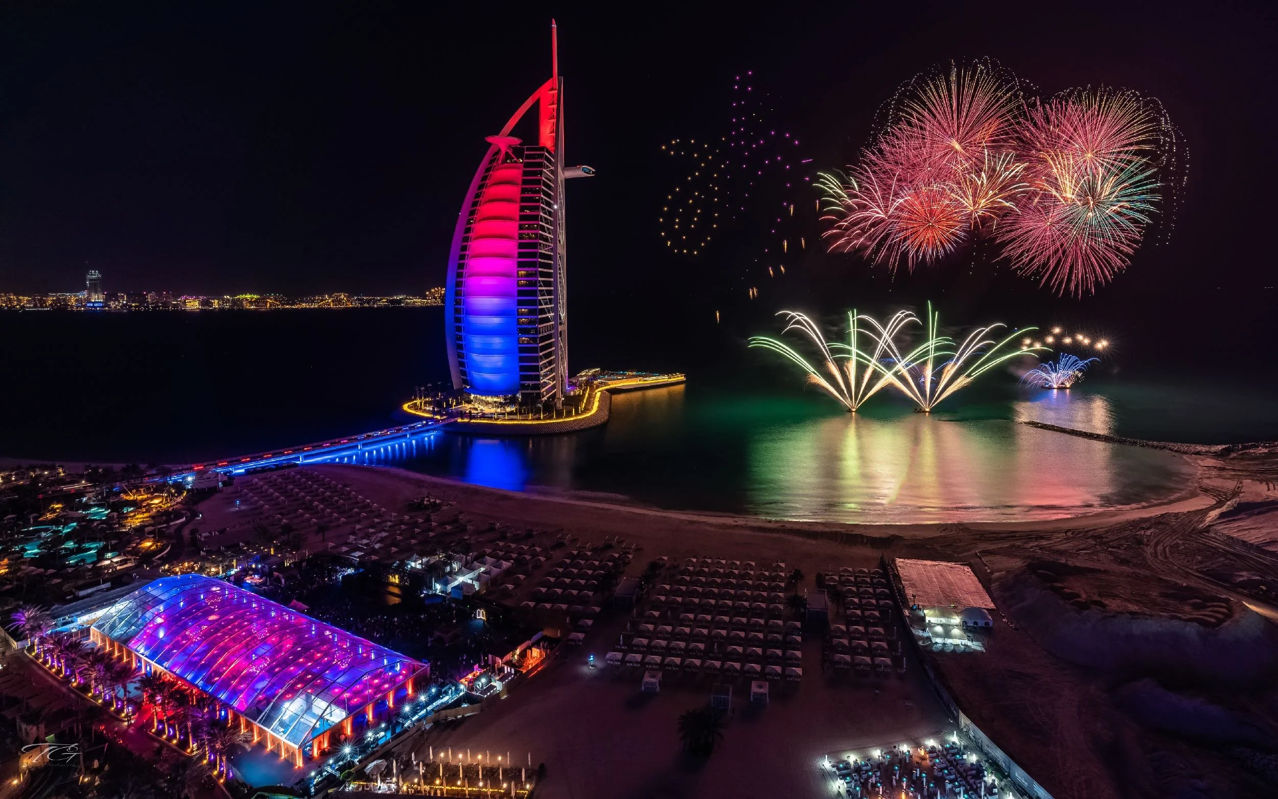 Nighttime fireworks display over the Burj Al Arab hotel in Dubai, illuminated with colorful lights, with a lighted dock and beach area in the foreground.