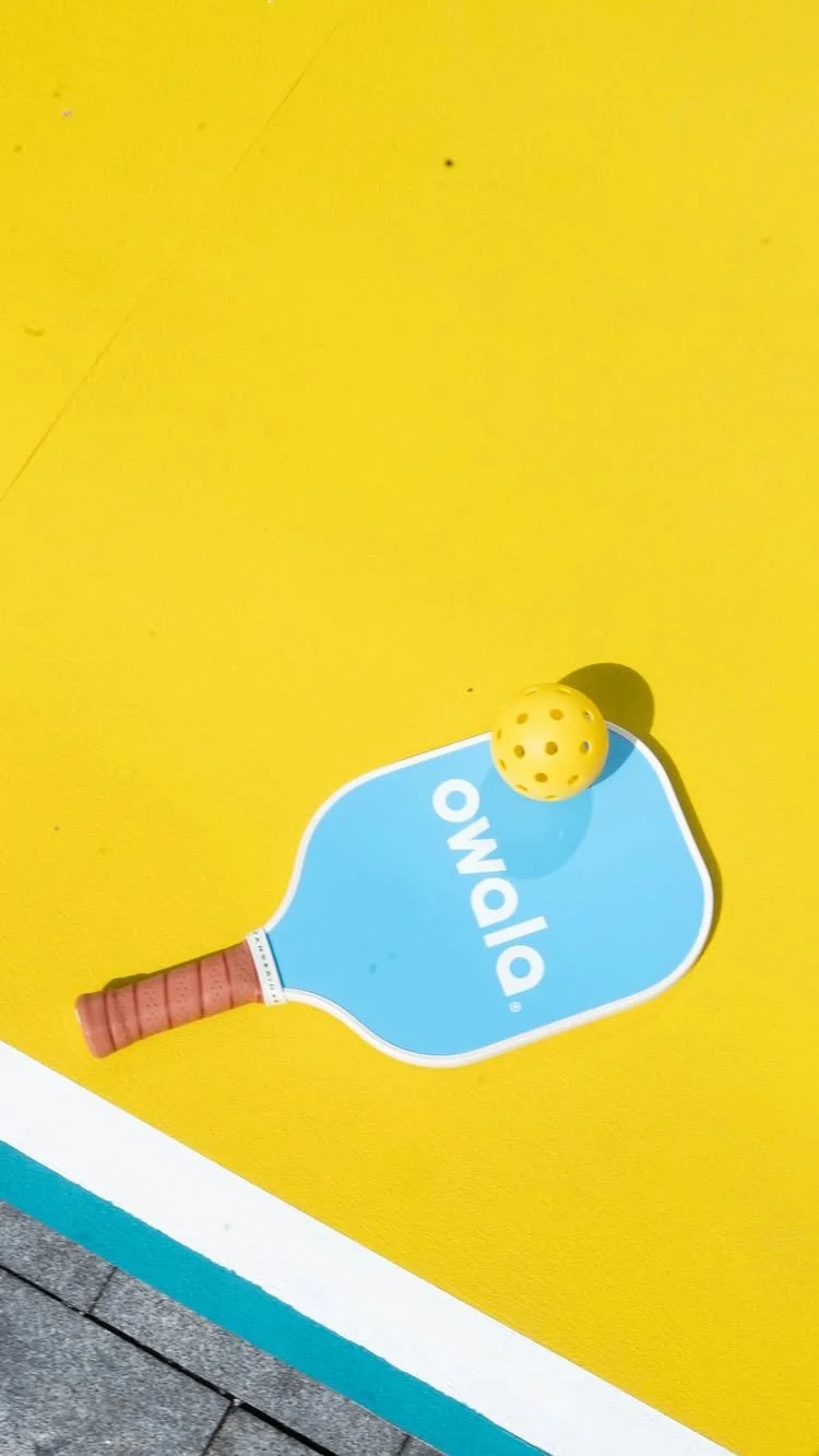 A blue paddle with the word "Olavo" written on it, a yellow ball with holes, and a pink handle on a yellow surface.