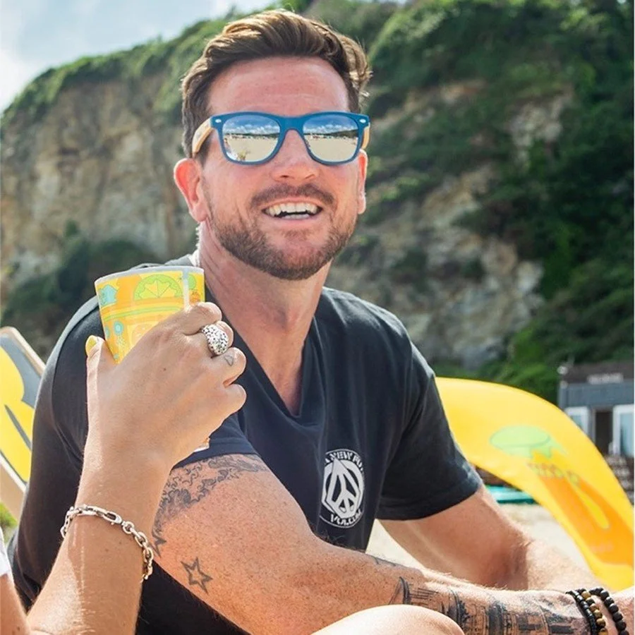 A young man in a gray polo shirt and beige pants sitting on a wooden bench against a beige wall, casting a shadow.