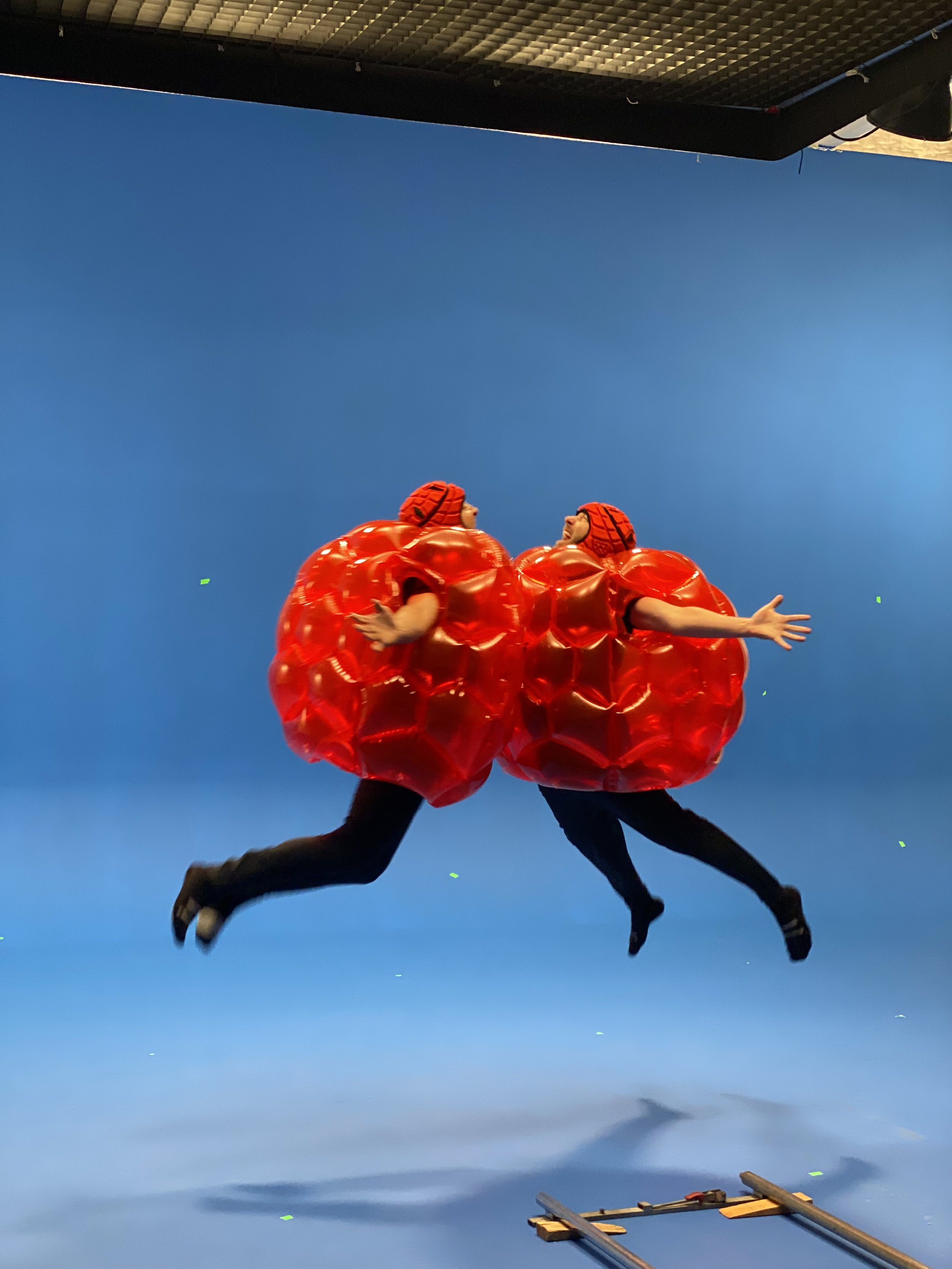 Two performers in red, inflatable costumes resembling tomatoes, jumping in front of a blue screen during a stage or film shoot.