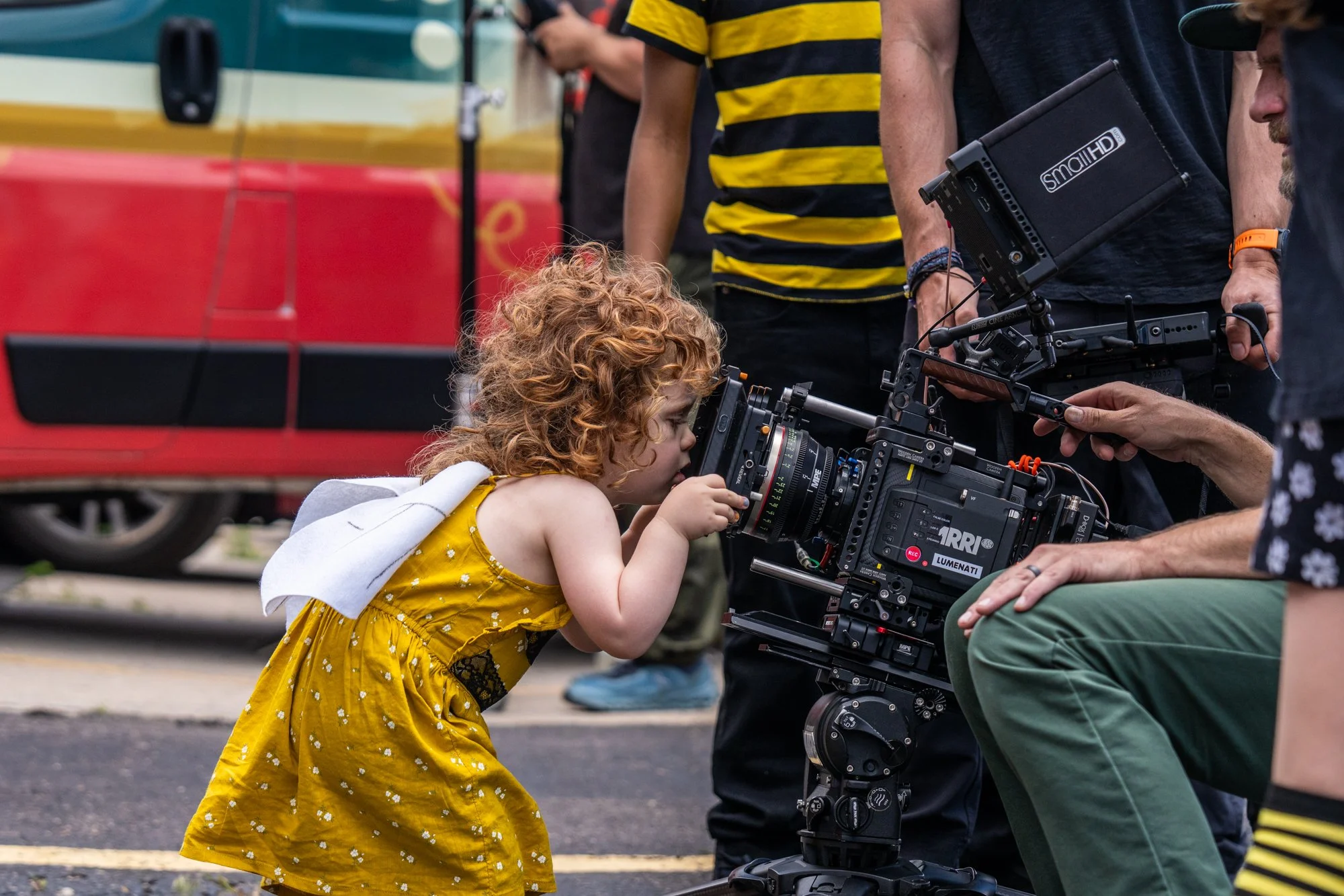 A young girl with curly red hair wearing a yellow dress looks through a camera mounted on a film set surrounded by crew members.