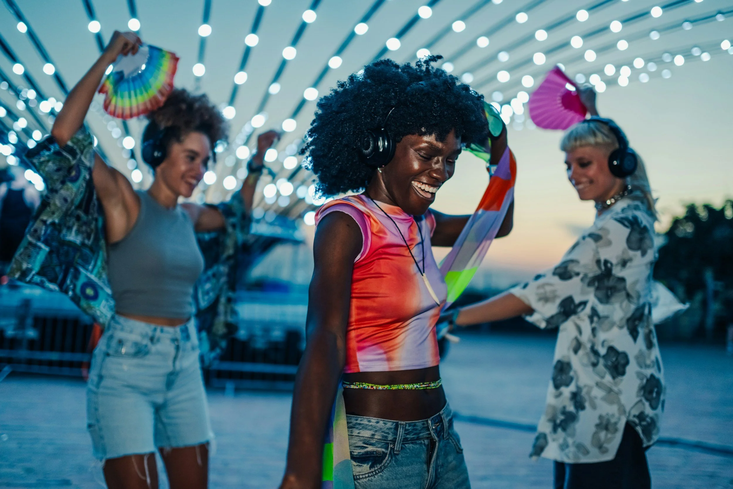 Group of young women dancing and enjoying music at an outdoor event during sunset, with string lights overhead.