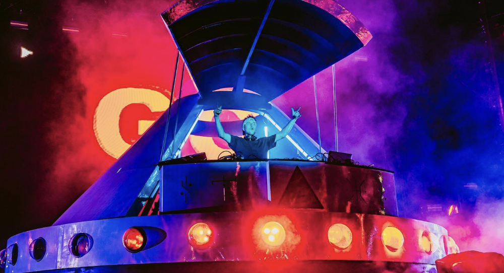 A DJ performs at a concert stage illuminated with vibrant purple and red lights, with haze and lighting effects in the background.