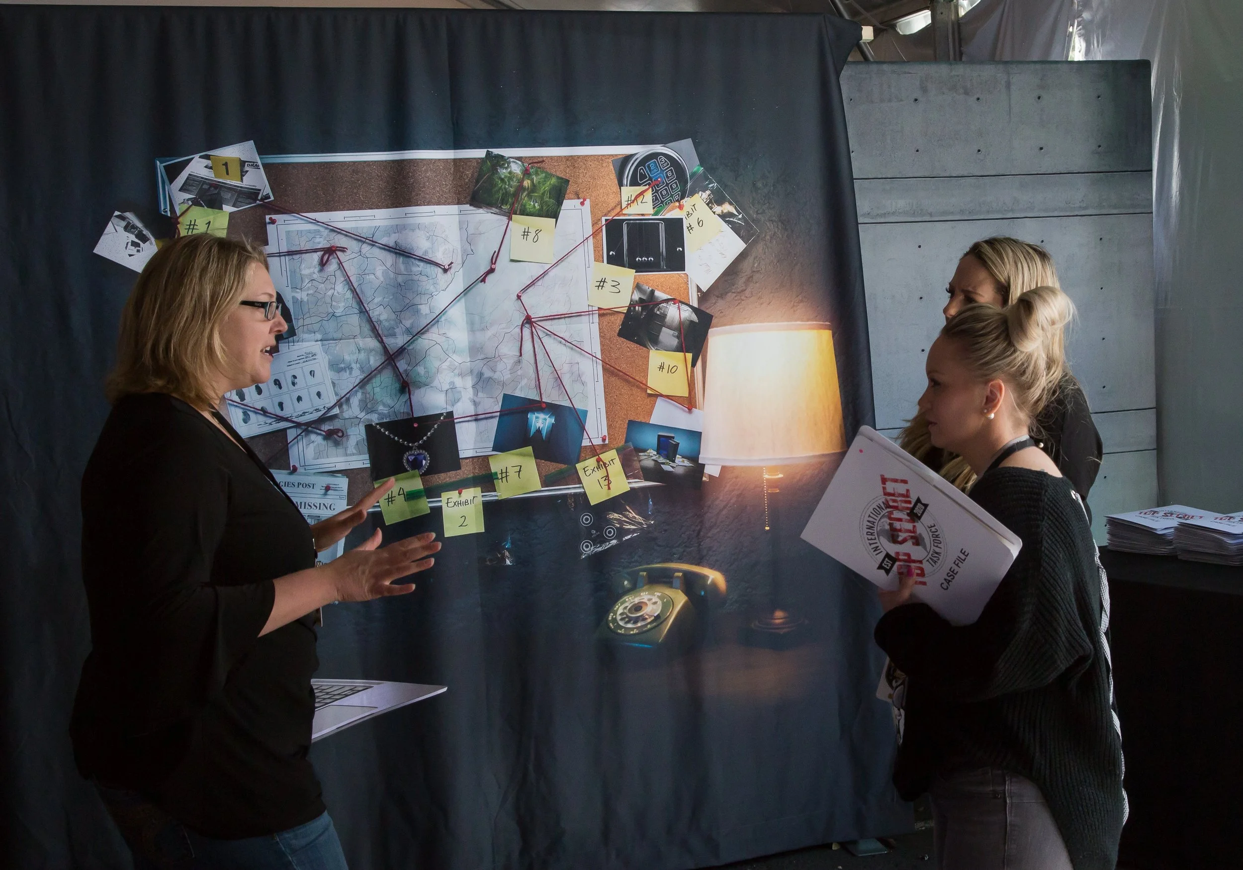 Three women discussing a crime investigation board with photos, notes, and maps, and a pile of case files on a table.