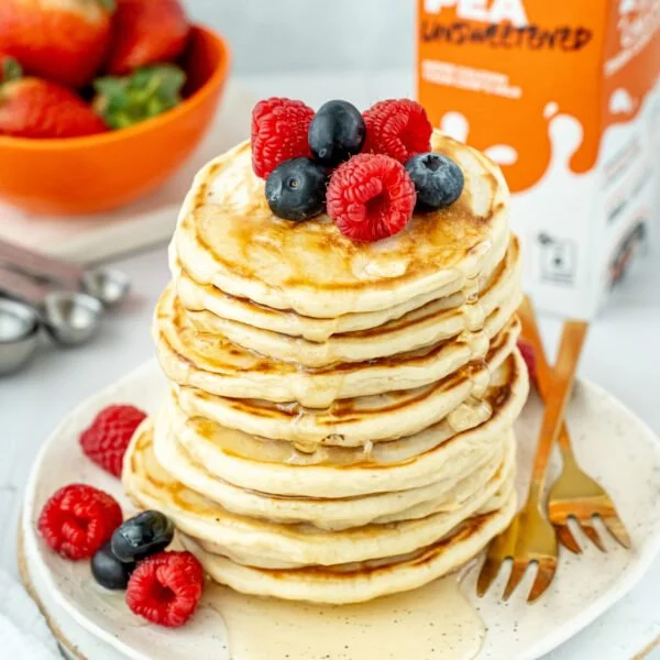 Stack of pancakes topped with mixed berries and syrup on a white plate, with a bowl of strawberries in the background.