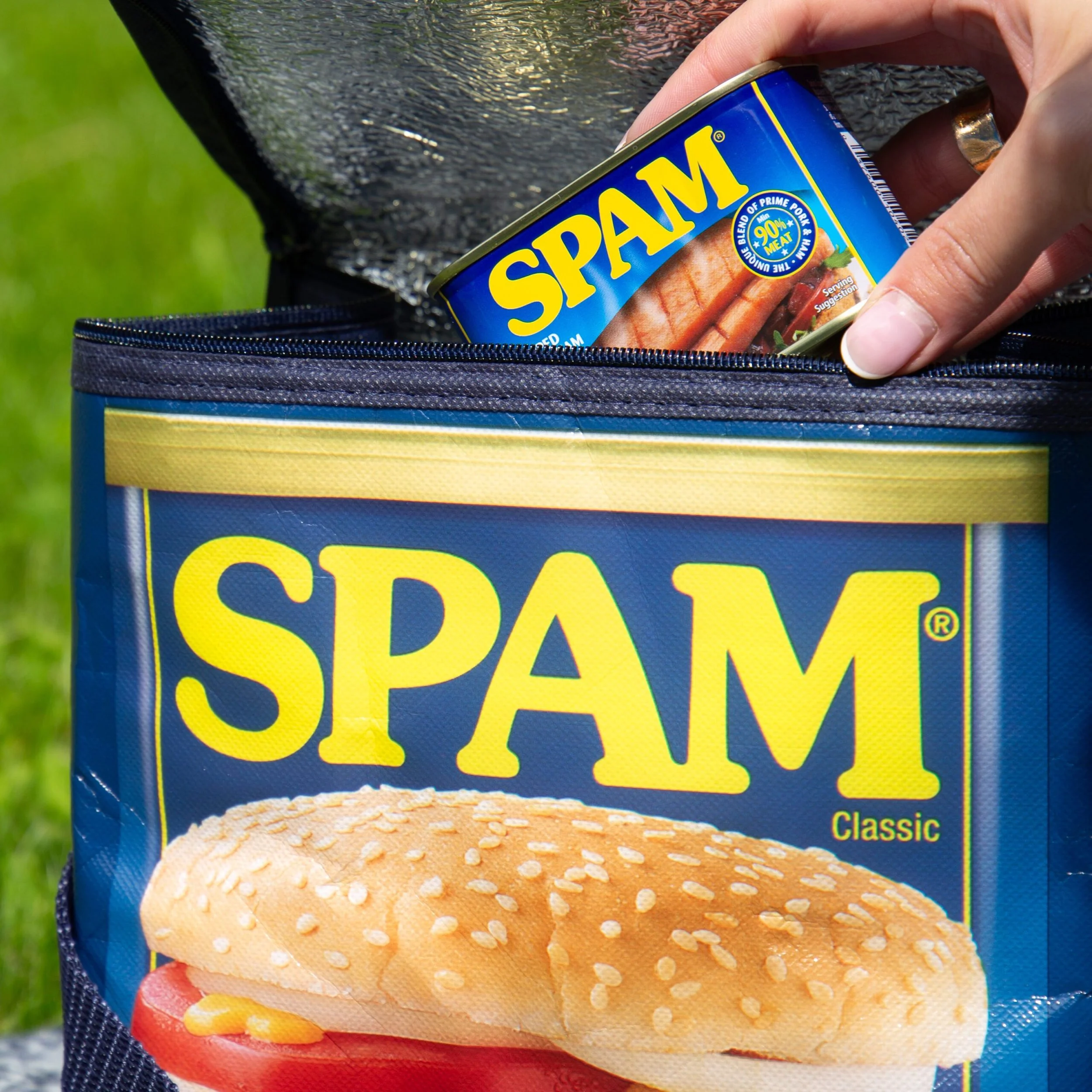 A hand placing a can of SPAM into a zippered bag with a large SPAM wrapper on it, outdoors on grass.
