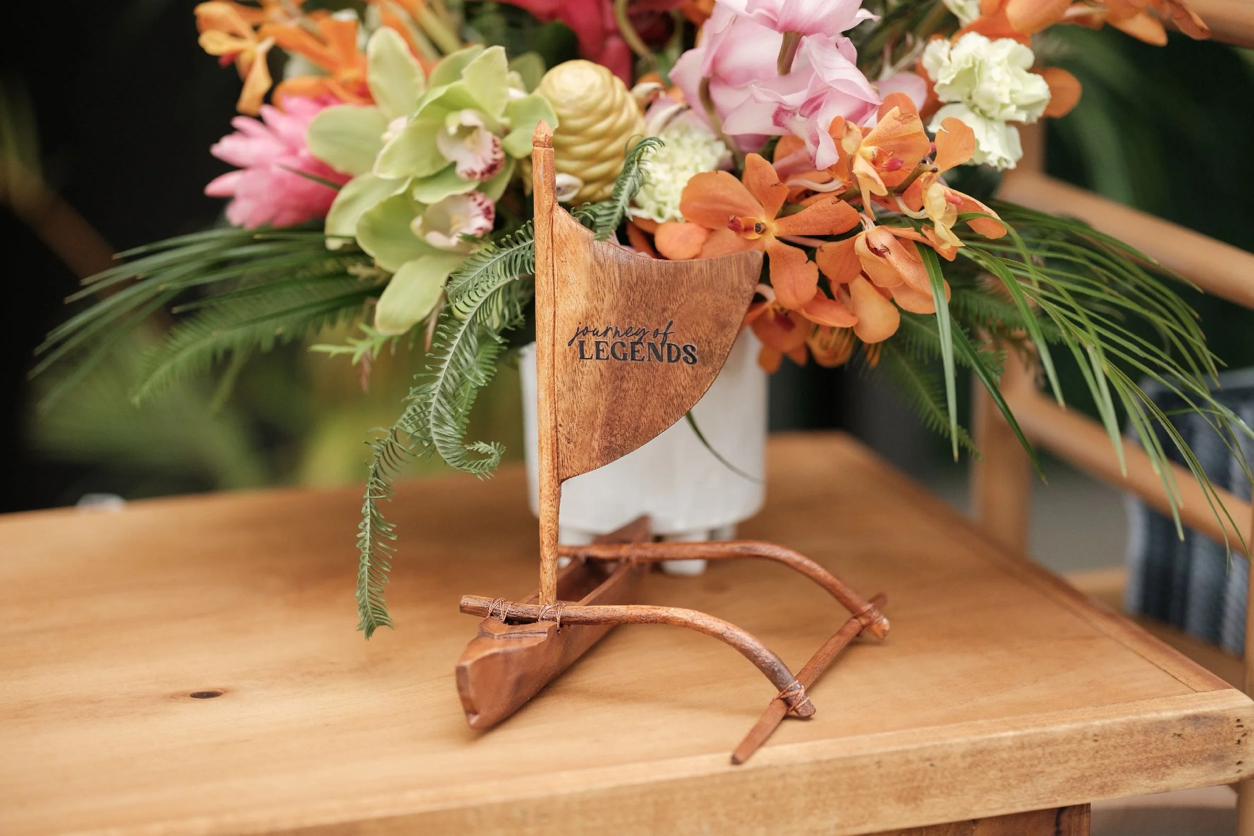 A small wooden boat with a flag reading 'journey of LEGENDS,' supporting a white flower vase with a colorful bouquet of pink, orange, and white flowers along with green fern leaves, on a wooden table.