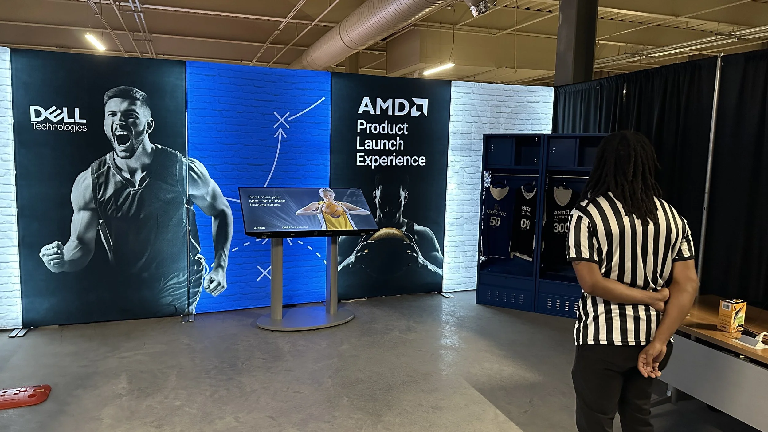 An AMD product launch experience booth with a large banner featuring a black and white image of a cheering athlete, and a person wearing a black and white striped shirt standing in front of lockers with AMD-branded merchandise.