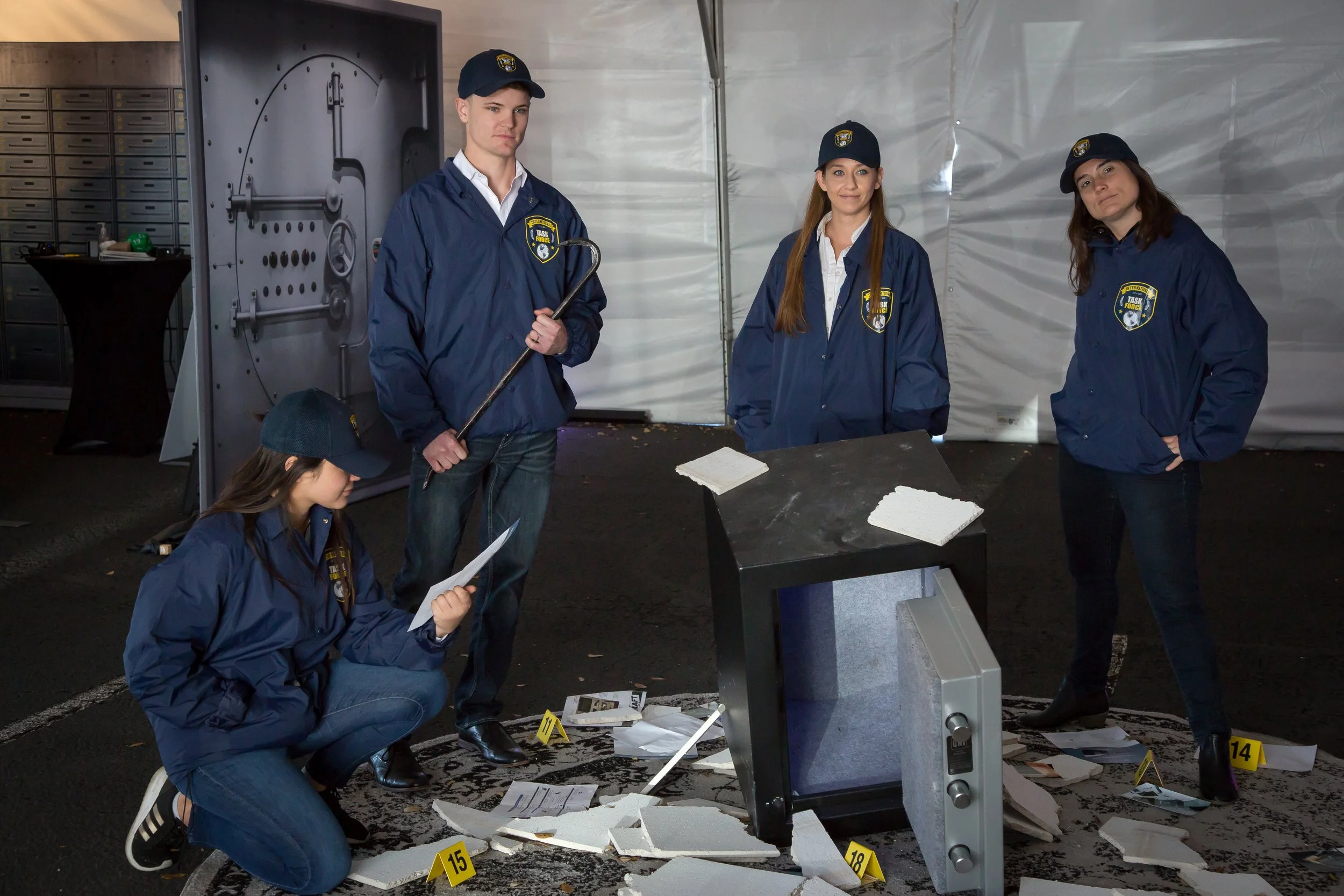 Five young crime scene investigators in police jackets and hats examine a broken television and scattered papers at a crime scene