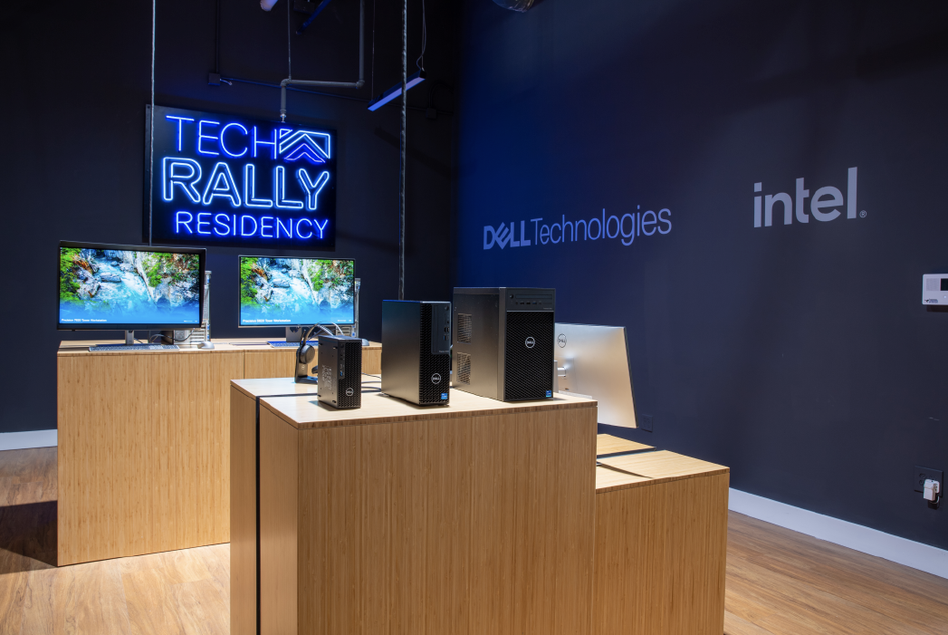 Tech Rally Residency event showcasing computer hardware, with Dell and Intel logos on the wall and two monitors displaying scenic images.
