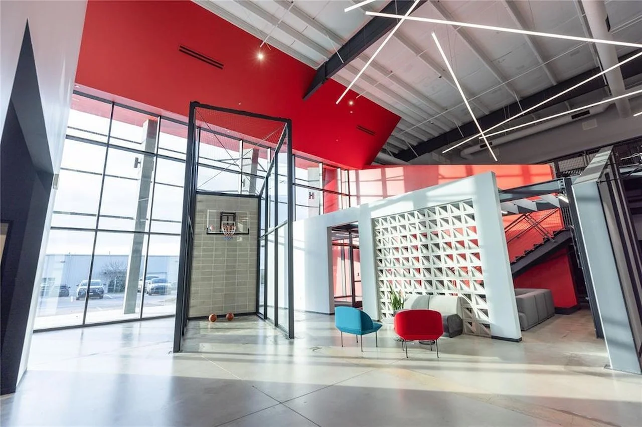 Modern interior space with basketball hoop and two basketballs, glass walls, red accents, and seating area with colorful chairs and a plant.