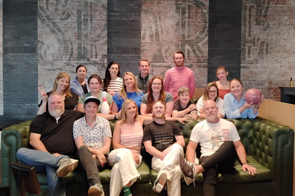 Group of 15 people gathered in a bar or game room, some sitting on a green leather sectional sofa and others standing behind, with a brick wall background and a bowling ball held by one woman.