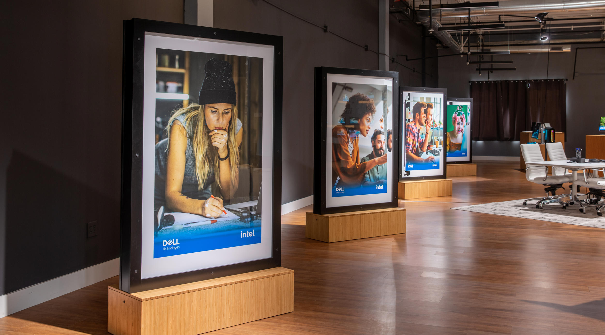 Series of four illuminated digital advertising displays featuring diverse young people in various activities, in a modern office space with wooden floors and white chairs.