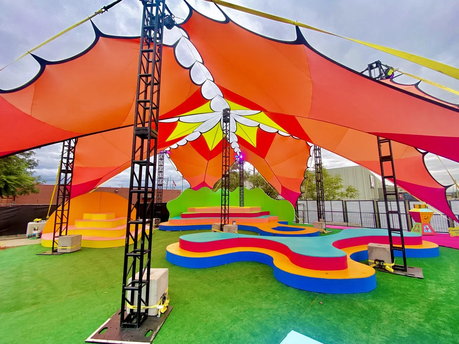 Colorful outdoor stage with a rainbow-colored canopy and stepping platforms, set up on a grassy area, with metal support structures and a bright sky in the background.