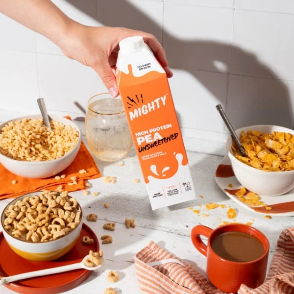 A hand holding a carton of Mighty High Protein Unsweetened milk over a table with cereal bowls, a glass of water, a cup of coffee, and a striped cloth.