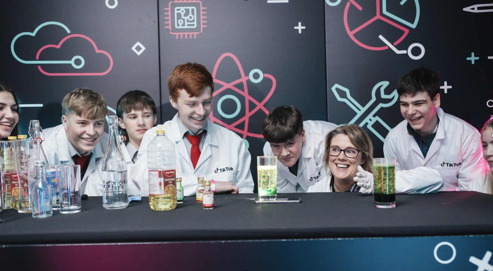 Group of children and an adult engaged in a science experiment with glowing liquids in beakers at a science demonstration or workshop, with science-themed wall art behind them.
