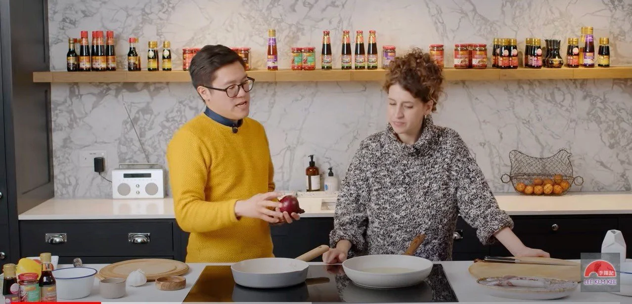 Two women preparing food in a modern kitchen with a marble backsplash. One woman is holding a red onion and wearing glasses and a yellow sweater. The other woman has curly hair, wears a patterned sweater, and is looking down. There are ingredients and kitchen utensils on the counter, including jars, a cutting board, and a fish.
