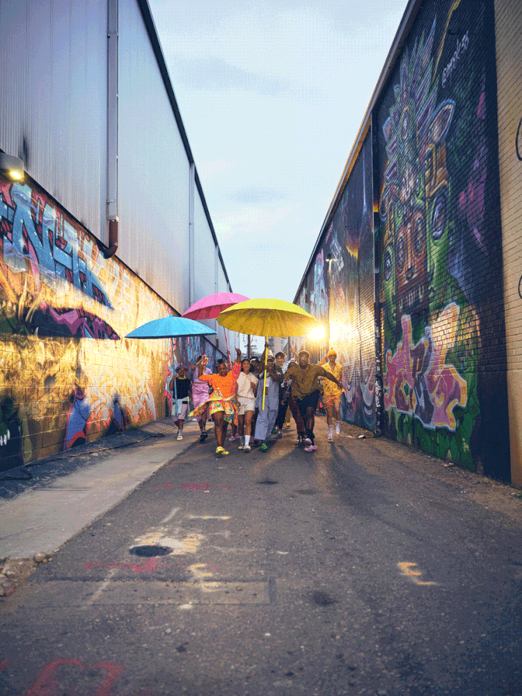 A group of people walking through a graffiti-covered alley at sunset, holding colorful umbrellas.