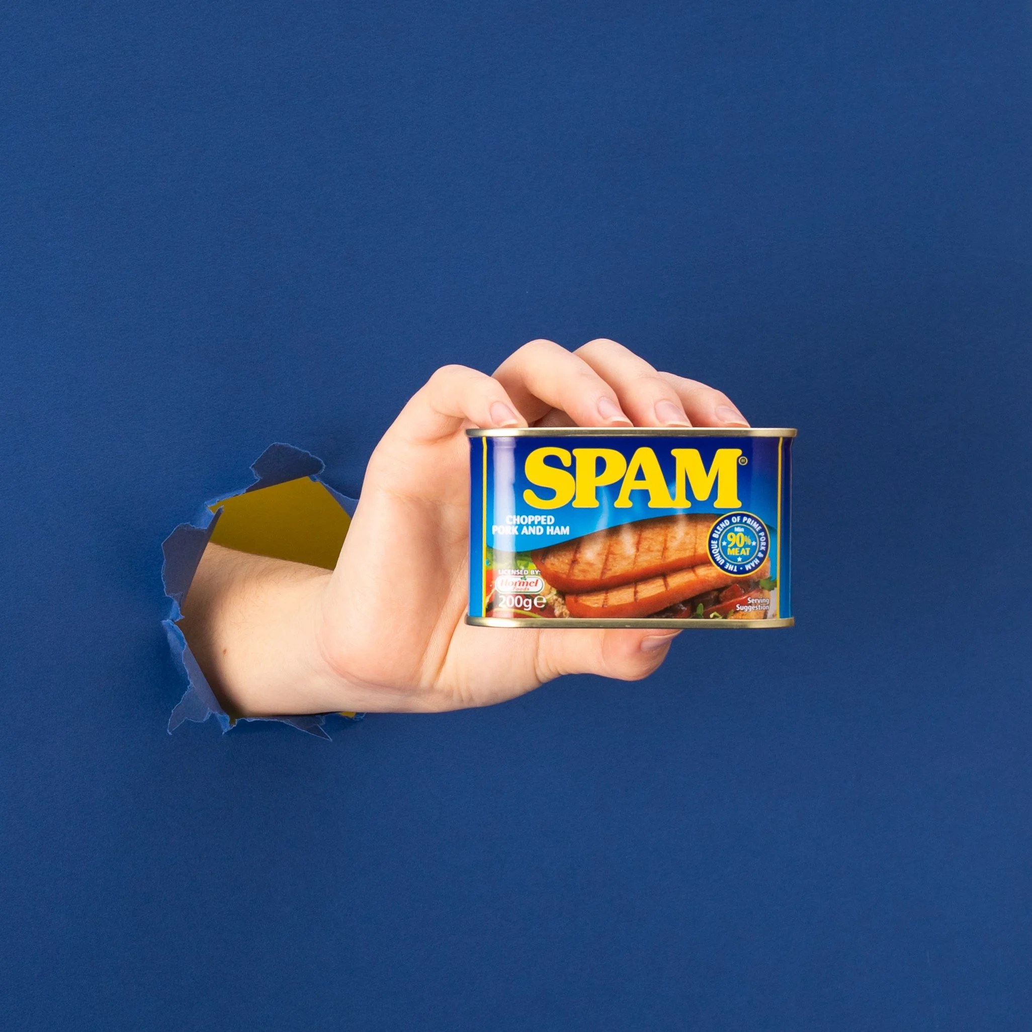 Hand coming through a hole in a blue background, holding a can of Spam with the label showing sliced pork and ham.