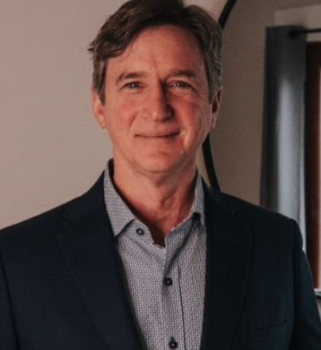 A middle-aged man with light brown hair, wearing a navy blazer over a light-colored dress shirt, standing indoors with a neutral background and a window with a blind.
