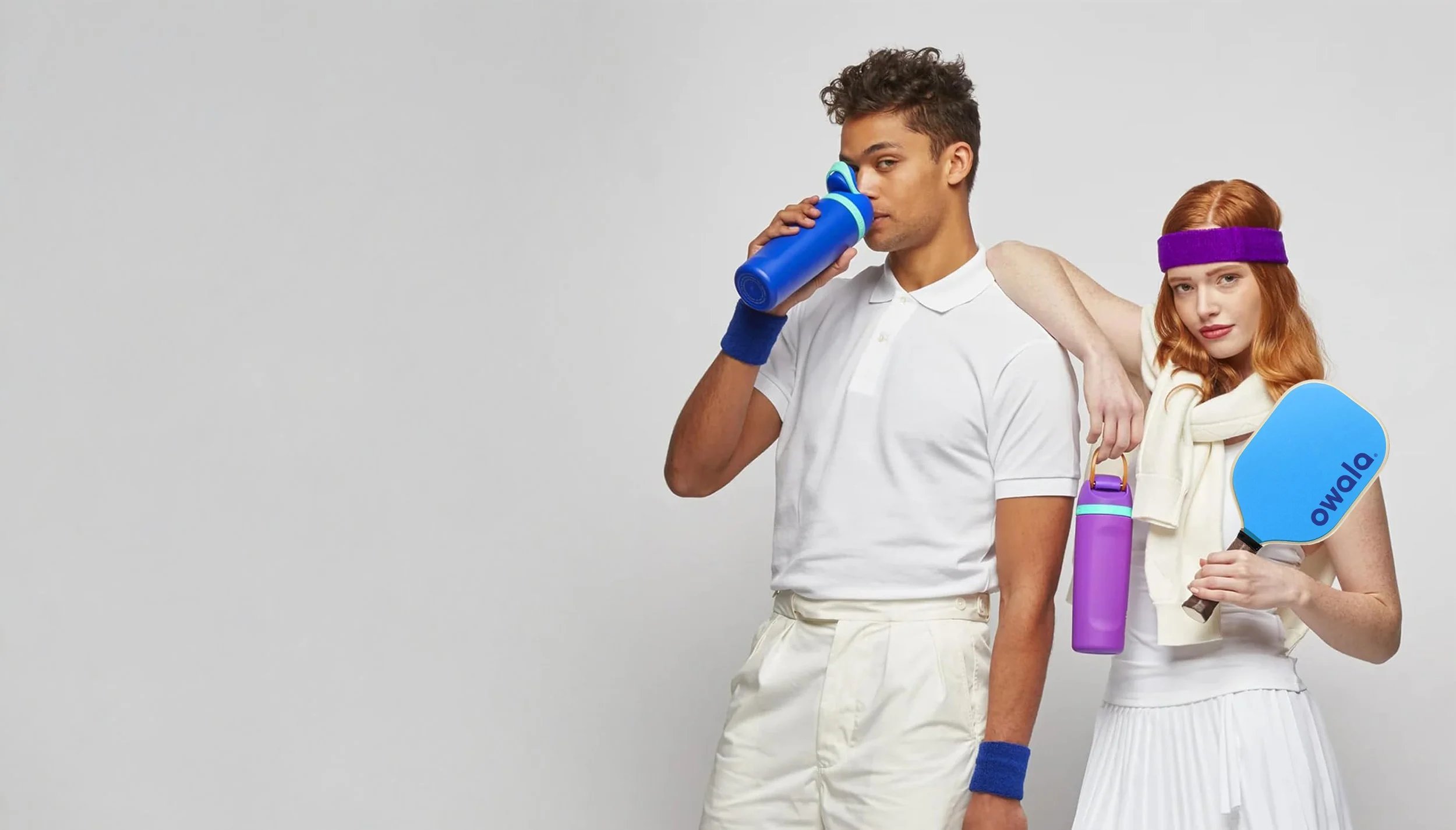 A man and a woman dressed in retro sportswear holding sports accessories, with the man drinking from a water bottle and the woman holding a table tennis paddle and a purple water bottle, standing against a plain light gray background.