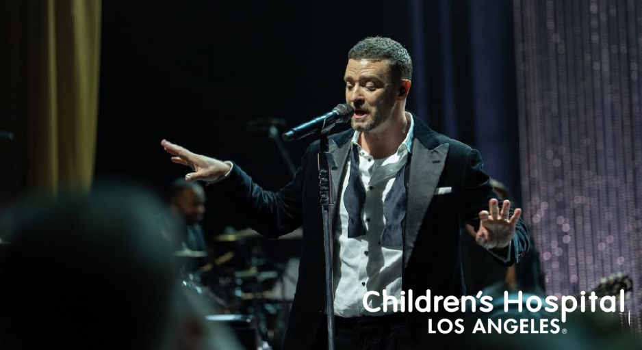 Man performing on stage in front of microphone with a band in the background, at a Children's Hospital event in Los Angeles.