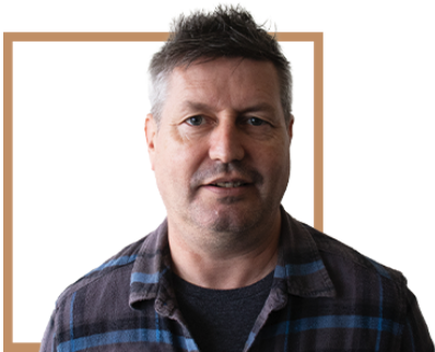 Portrait of a man with short, dark, and graying hair, wearing a dark plaid shirt, against a plain background with a light brown border.