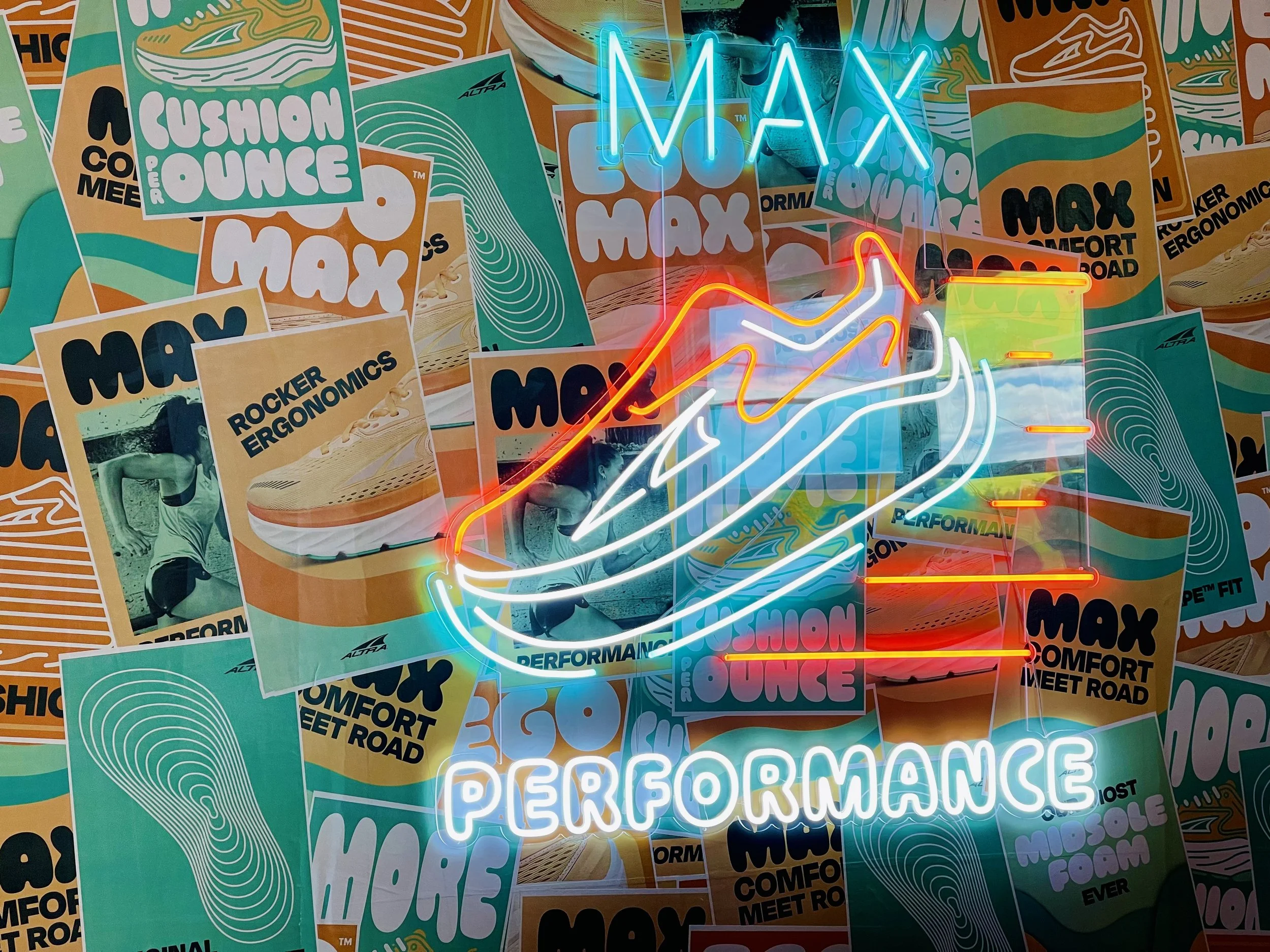 A colorful wall decorated with numerous Max branding posters, featuring slogans such as "Cushion Ounce" and "Rocker Ergonomics," along with a neon sign displaying an abstract shoe design and the words "Max Performance."