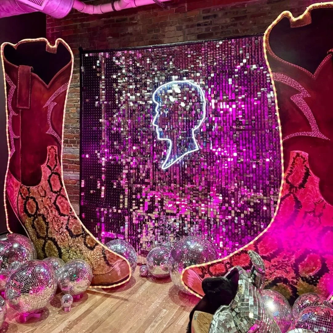 Decorative display featuring a neon outline of a human head in profile, surrounded by numerous disco balls, with pink and purple lighting effects, large colorful fabric panels on either side, and a brick wall in the background.