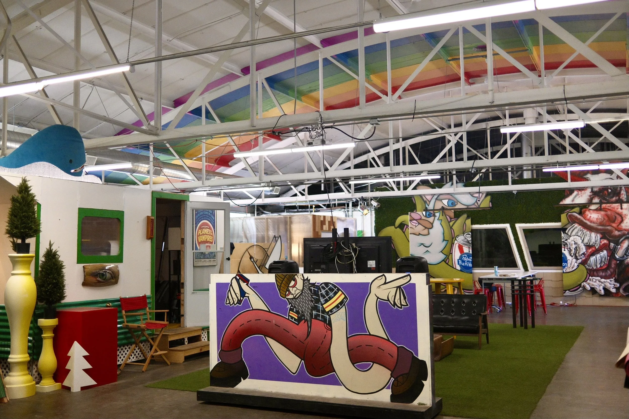 An indoor space with colorful graffiti art and decorations, including cartoon characters and furniture, and a DJ setup in the center.