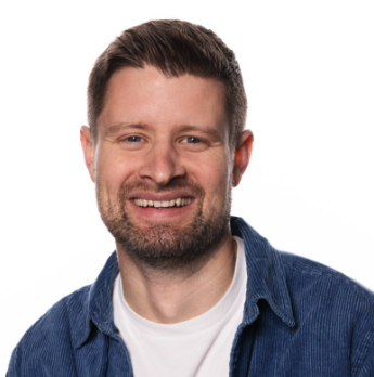 A smiling man with a beard and short dark hair, wearing a white shirt and blue denim jacket.