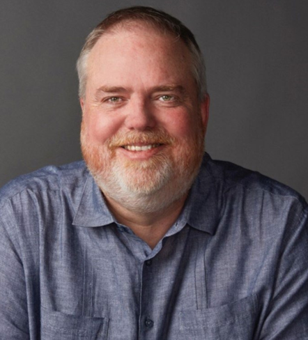 A smiling man with short gray hair and a beard, wearing a blue collared shirt, against a plain gray background.