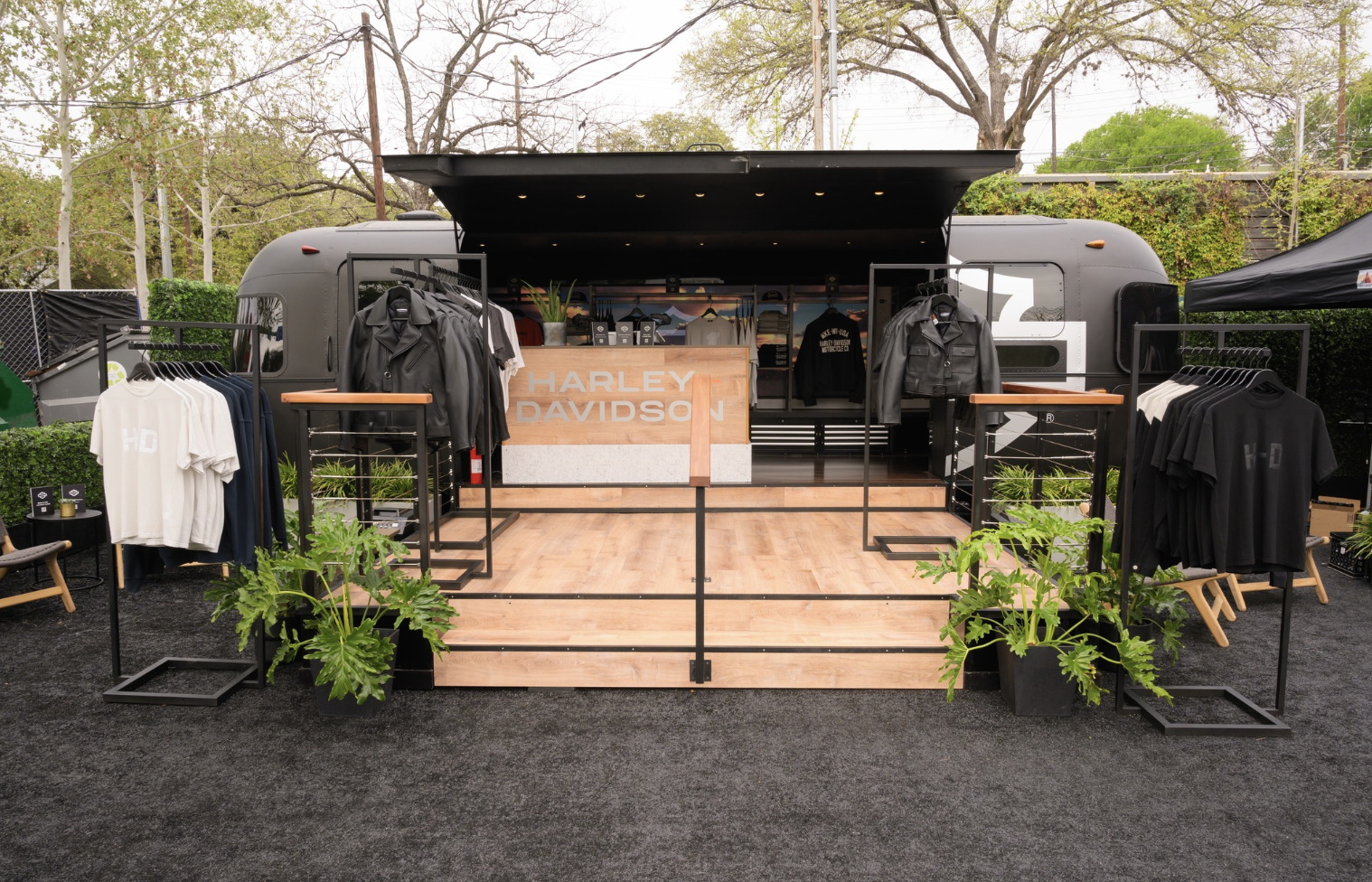 Outdoor Harley Davidson merchandise display inside a trailer with clothing racks, potted plants, and a wooden stage for sales.