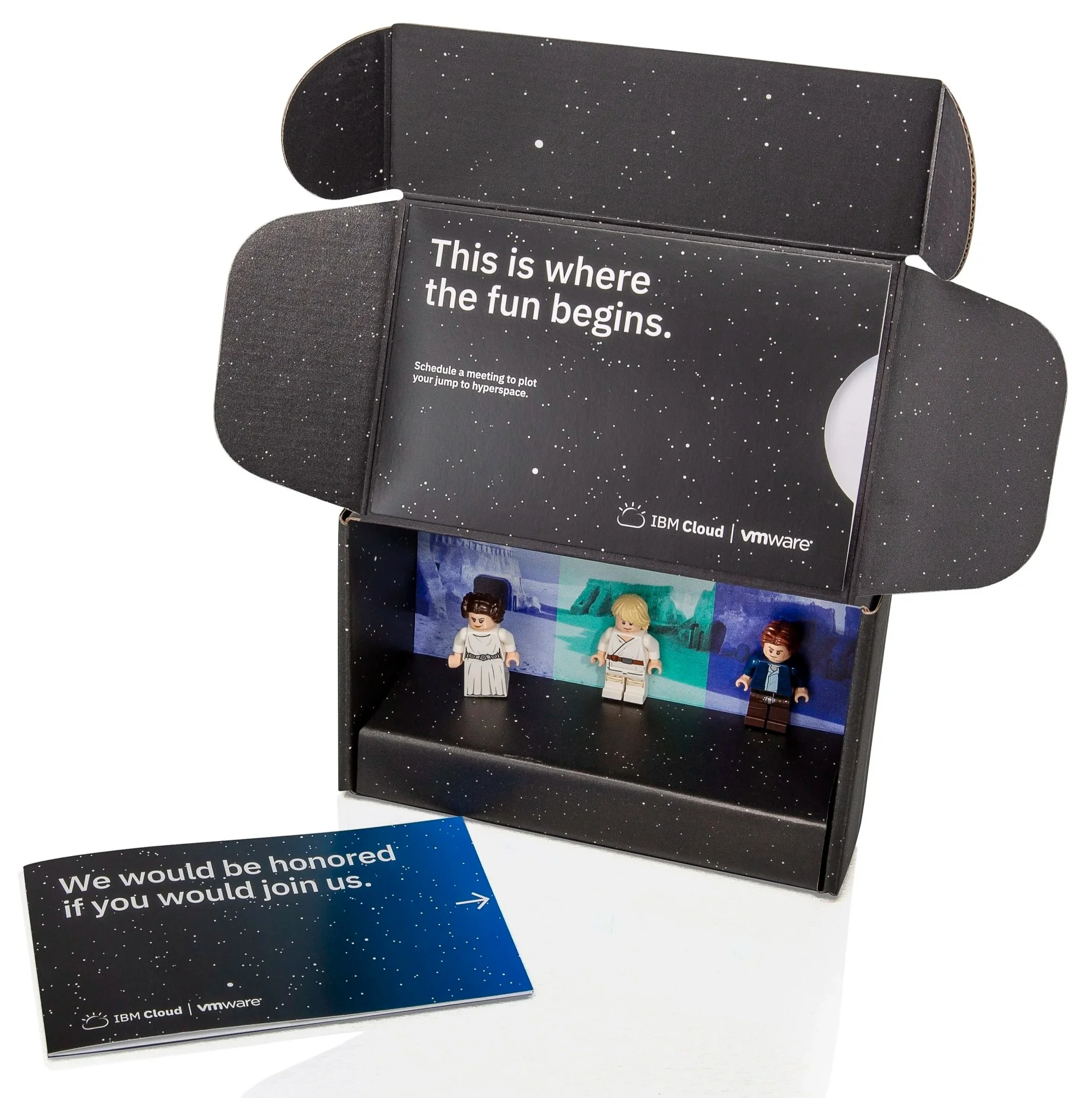A black box with a starry night sky design contains three LEGO minifigures standing on a platform with a space-themed background. The box has the text 'This is where the fun begins' and a message to schedule a meeting. An invitation card beside the box reads, 'We would be honored if you would join us.'