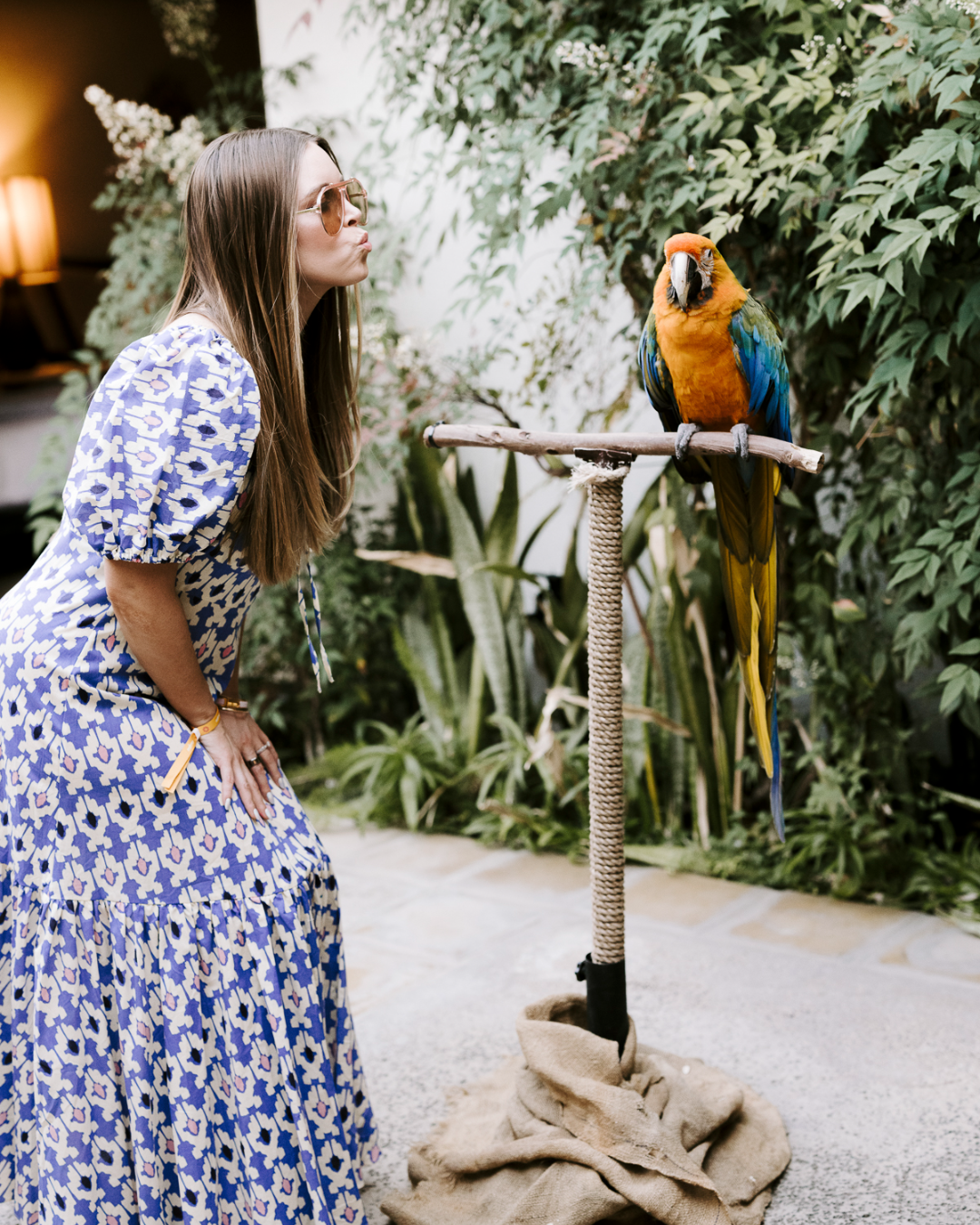 A woman in a patterned dress and sunglasses blowing a kiss toward a colorful macaw perched on a stand surrounded by lush green indoor plants.