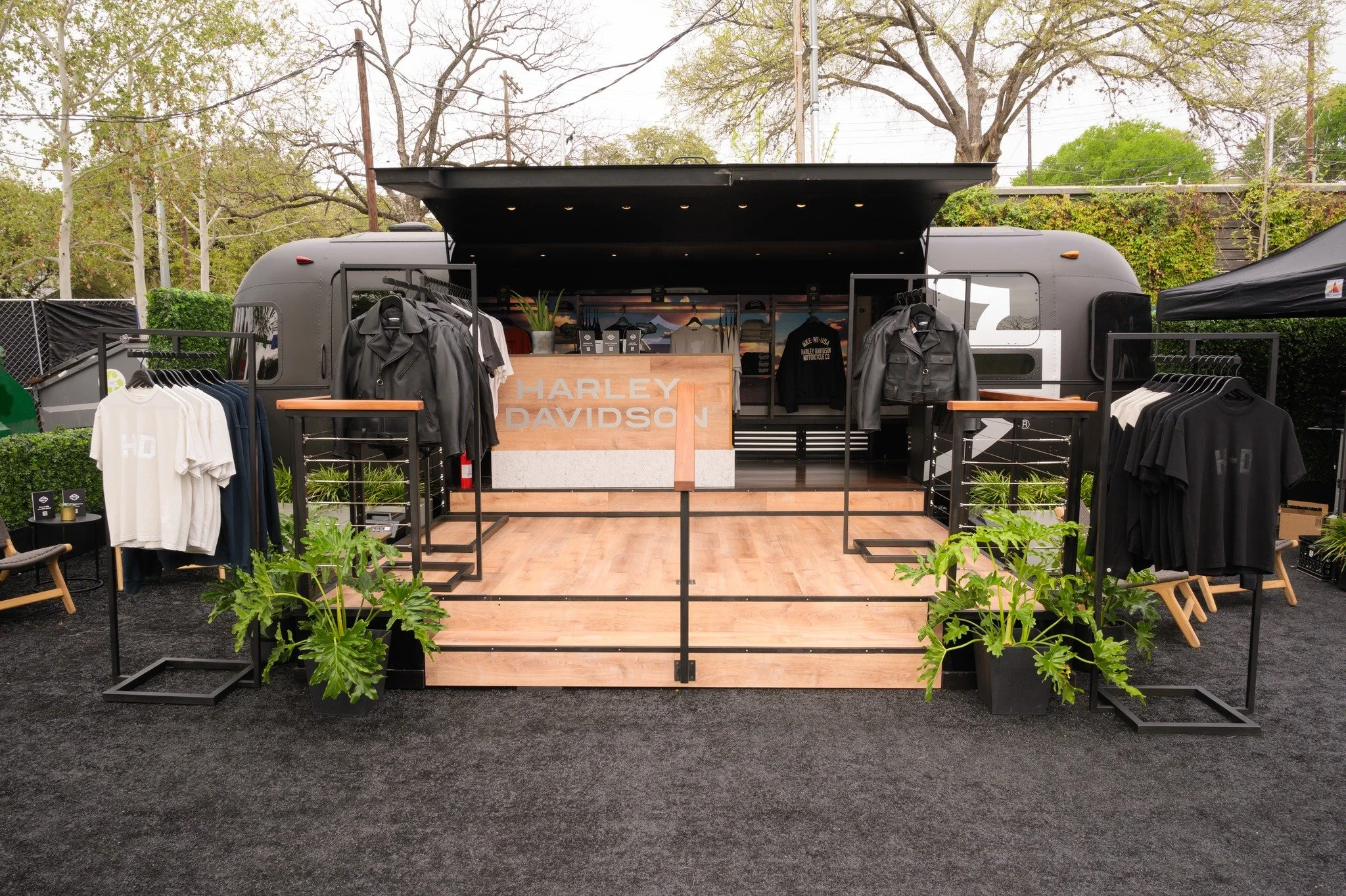 A mobile Harley Davidson retail display inside a black trailer with clothing items such as t-shirts, jackets, and hats, set up on a wooden platform with plants and black carpeted flooring outside.