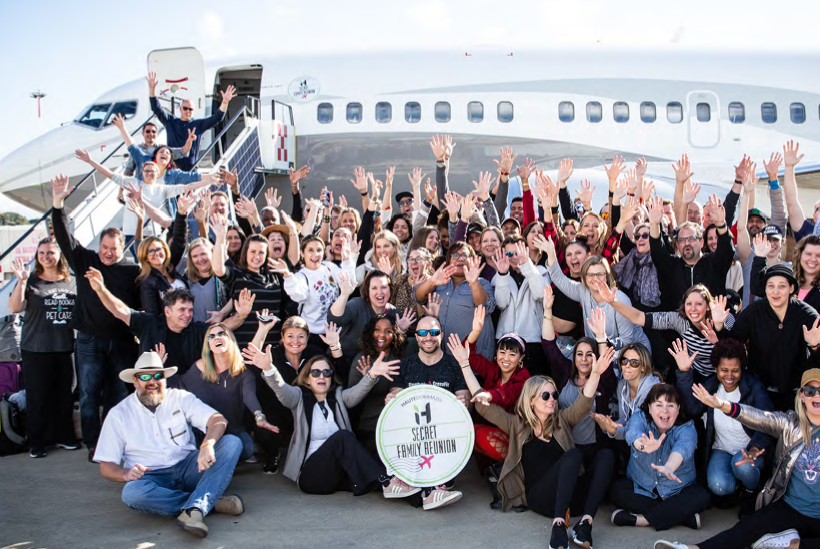 Large group of people posing with raised hands in front of an airplane during a family reunion, with some seated on the ground and others standing on stairs leading to the aircraft.